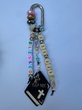 The design includes cream letter blocks spelling 'I I I I', a silver-tone carabiner clip, assorted colorful beads, a small 'BLESSED' letter cube phrase, a black book charm with a white cross and the words 'TAP ME', and a silver cross charm.